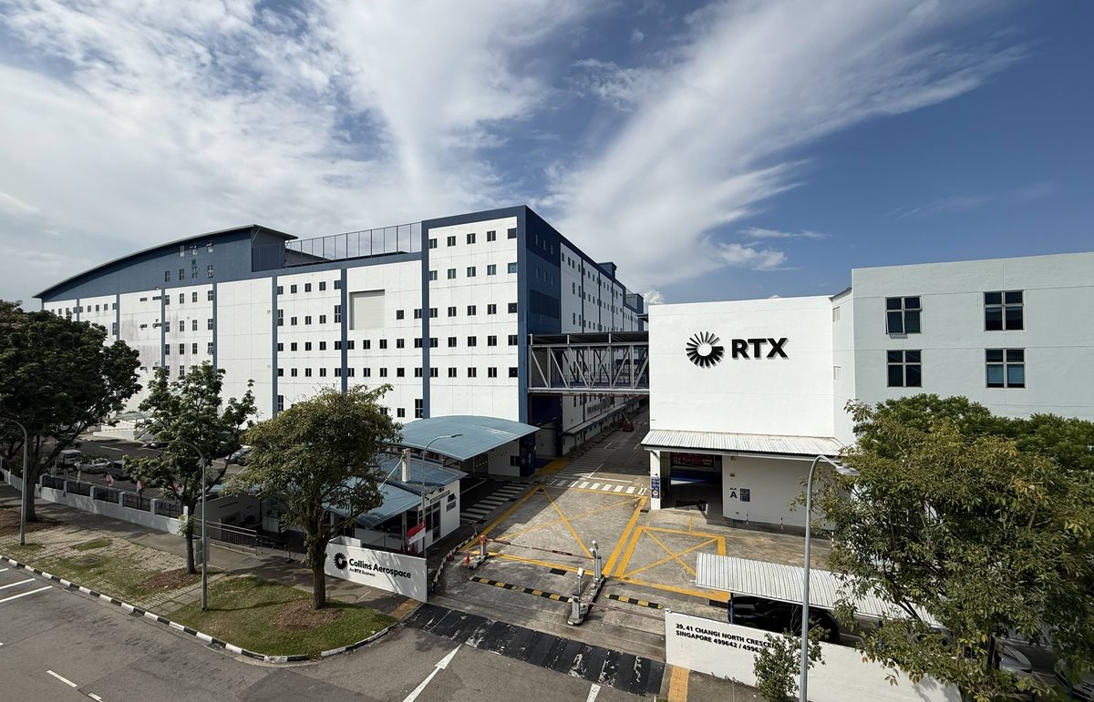 GASCA Site Photo Hi Res v2 1jpg Wide angle view of an industrial campus white and blue buildings with an RTX sign connected by a pedestrian bridge under a clear sky with trees and road lanes nearby