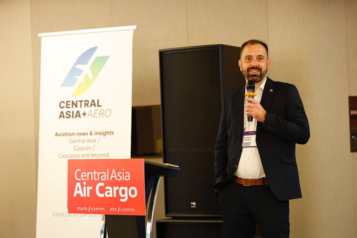 Gerhard Coetzee CAACS Man in a dark suit speaking into a microphone at a conference with a Central Asia Aero banner and a red Central Asia Air Cargo sign beside him
