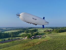 White HyLight airship gliding over green vineyards and rolling countryside with a blue tail in the distance to the right