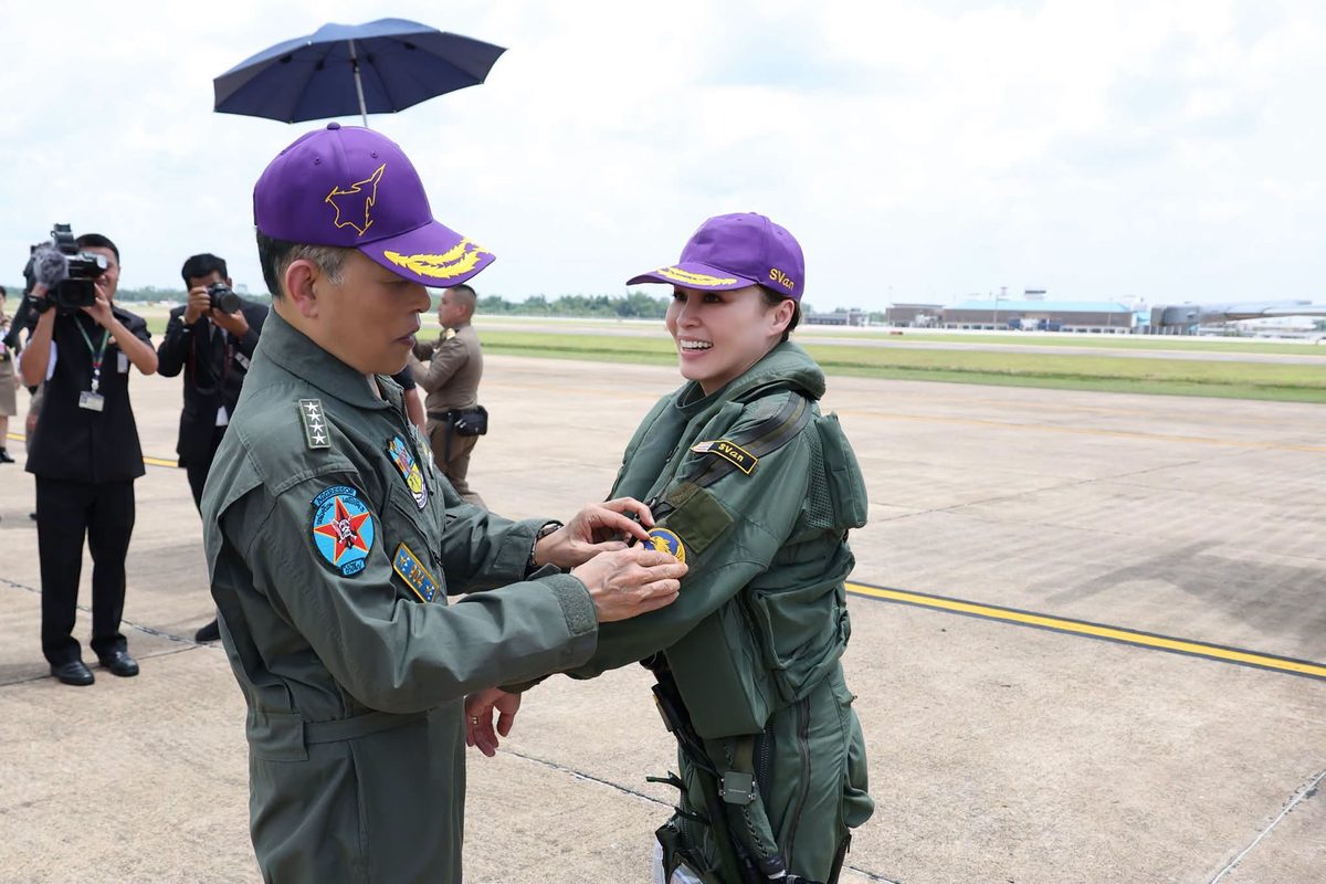 King Maha Vajiralongkorn presenting the Golden Gripen Order Patch to Queen Suthida after her solo flight Two military personnel on a runway exchange a medal during a ceremony with photographers filming in the background