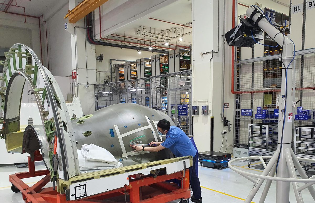 Nacelles MRO GASCA 4jpg A technician in a blue shirt and mask works on a large aircraft cockpit section mounted on a red and orange frame in an industrial workshop