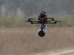 Quadcopter drone with a cylindrical payload hanging from a tether over a grassy field