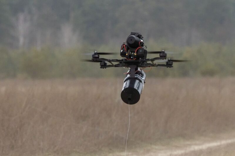 Quadcopter drone with a cylindrical payload hanging from a tether over a grassy field