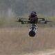 Quadcopter drone with a cylindrical payload hanging from a tether over a grassy field