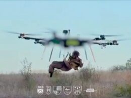 A soldier suspended in a harness beneath a large quadcopter drone hovering over a dry grassy field logos shown at the bottom
