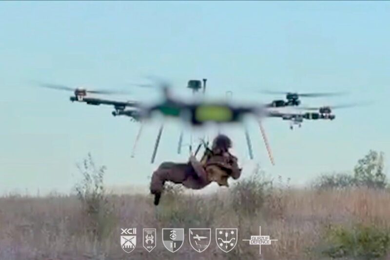 Recently Saved 1 of 1 10 A soldier suspended in a harness beneath a large quadcopter drone hovering over a dry grassy field logos shown at the bottom