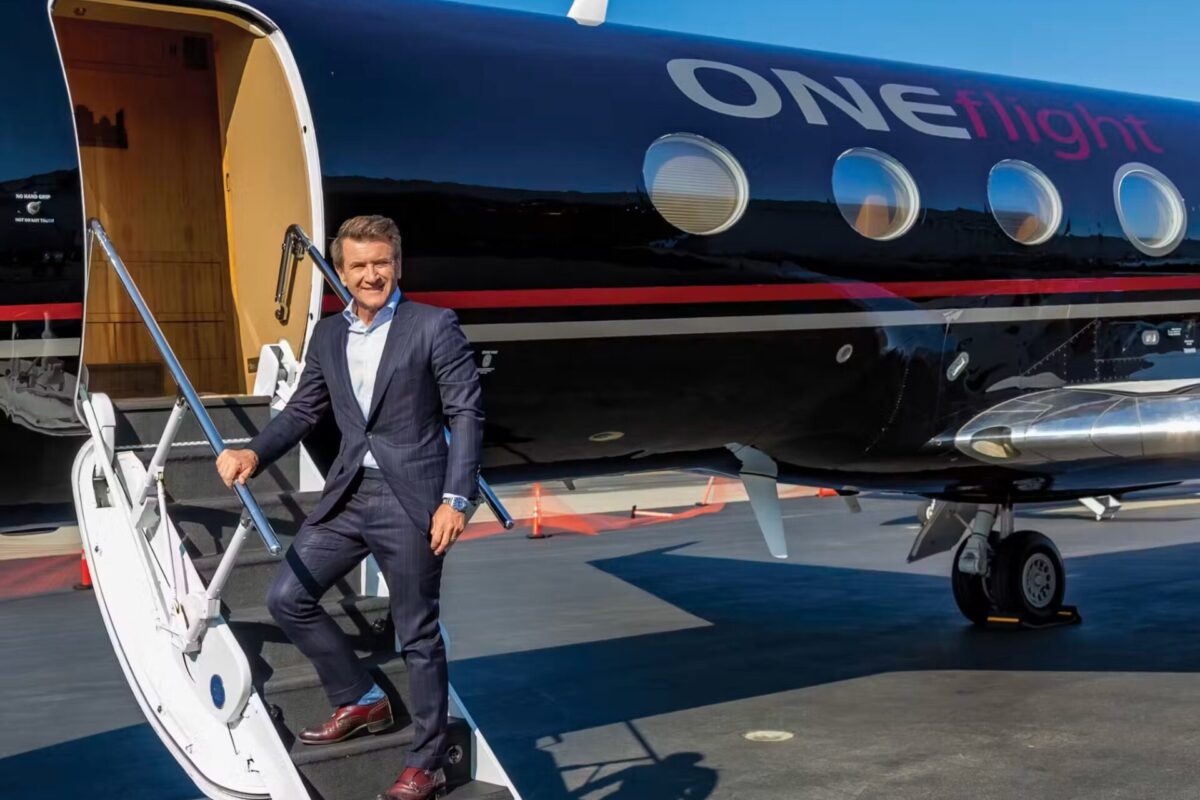 Man in a suit stepping down a white stairway from a dark blue private jet branded ONEflight on a sunny tarmac