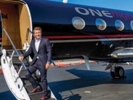 Man in a suit stepping down a white stairway from a dark blue private jet branded ONEflight on a sunny tarmac
