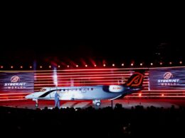 Airliner on a stage at night with red lights and large screens reading SYBERJET WORLD audience silhouettes in the foreground