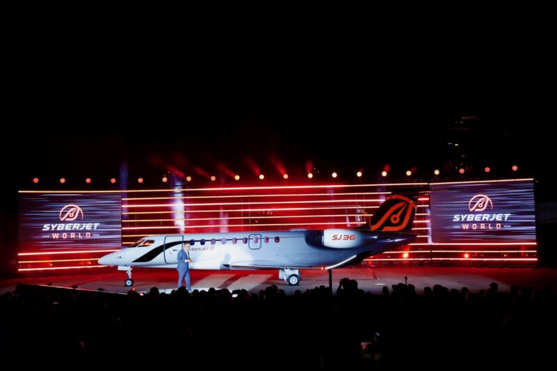 Airliner on a stage at night with red lights and large screens reading SYBERJET WORLD audience silhouettes in the foreground