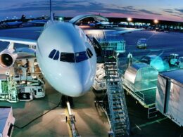 Front view of a white passenger aircraft at a gate with stairs and jet bridge at dusk with ground support equipment nearby