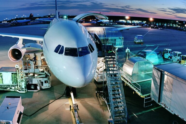 Front view of a white passenger aircraft at a gate with stairs and jet bridge at dusk with ground support equipment nearby