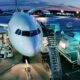 Front view of a white passenger aircraft at a gate with stairs and jet bridge at dusk with ground support equipment nearby