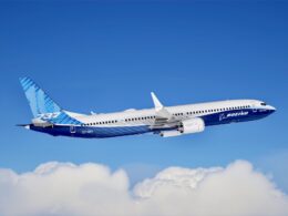 Blue and white Boeing 737 MAX airliner flying high above fluffy clouds against a clear blue sky