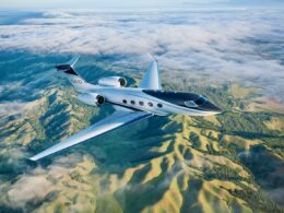White Gulfstream G400 private jet cruising over a green valley with clouds below and distant hills