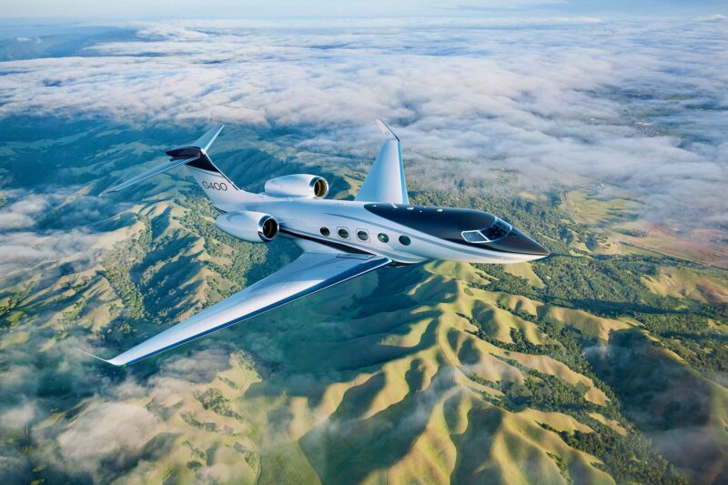 White Gulfstream G400 private jet cruising over a green valley with clouds below and distant hills