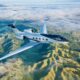 White Gulfstream G400 private jet cruising over a green valley with clouds below and distant hills