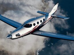 White and blue private jet with red stripe flying above clouds viewed from the front left