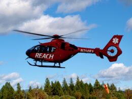 Red emergency medical helicopter in flight over a forest under a blue sky with REACH branding on the side and a white star of life on the tail