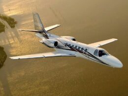 White private jet with black stripes flying over a sunlit body of water at sunset aerial view Citation Encore logo visible