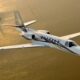 White private jet with black stripes flying over a sunlit body of water at sunset aerial view Citation Encore logo visible