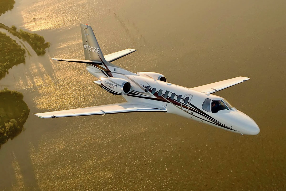 White private jet with black stripes flying over a sunlit body of water at sunset aerial view Citation Encore logo visible