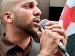 Close up of a man singing into a microphone at a public event with a red and white flag visible in the background