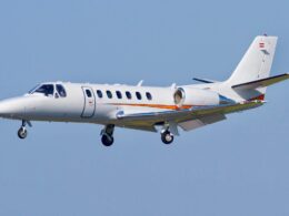 White business jet in flight against a clear blue sky with an orange stripe along the fuselage and gear extended for landingapproach tailswept