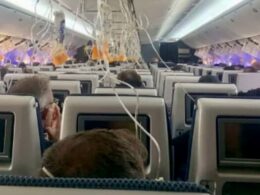 Inside an airplane cabin with ceiling panels open and many tangled wires hanging down displays screens on seatbacks are visible among the clutter