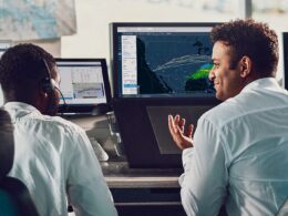 Two operators in a control room monitor a large digital map showing weather patterns and routes on a central screen one gesturing as they discuss data