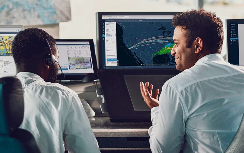 Recently Saved 1 of 1 34 Two operators in a control room monitor a large digital map showing weather patterns and routes on a central screen one gesturing as they discuss data