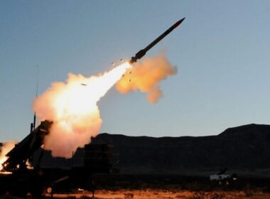Ground-based missile launcher fires a rocket, bright flame and smoke plume streaking into the sky with desert terrain in the background.