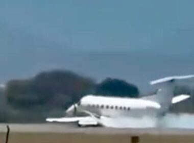 Small white jet parked on a runway with trees and a blue sky in the background.