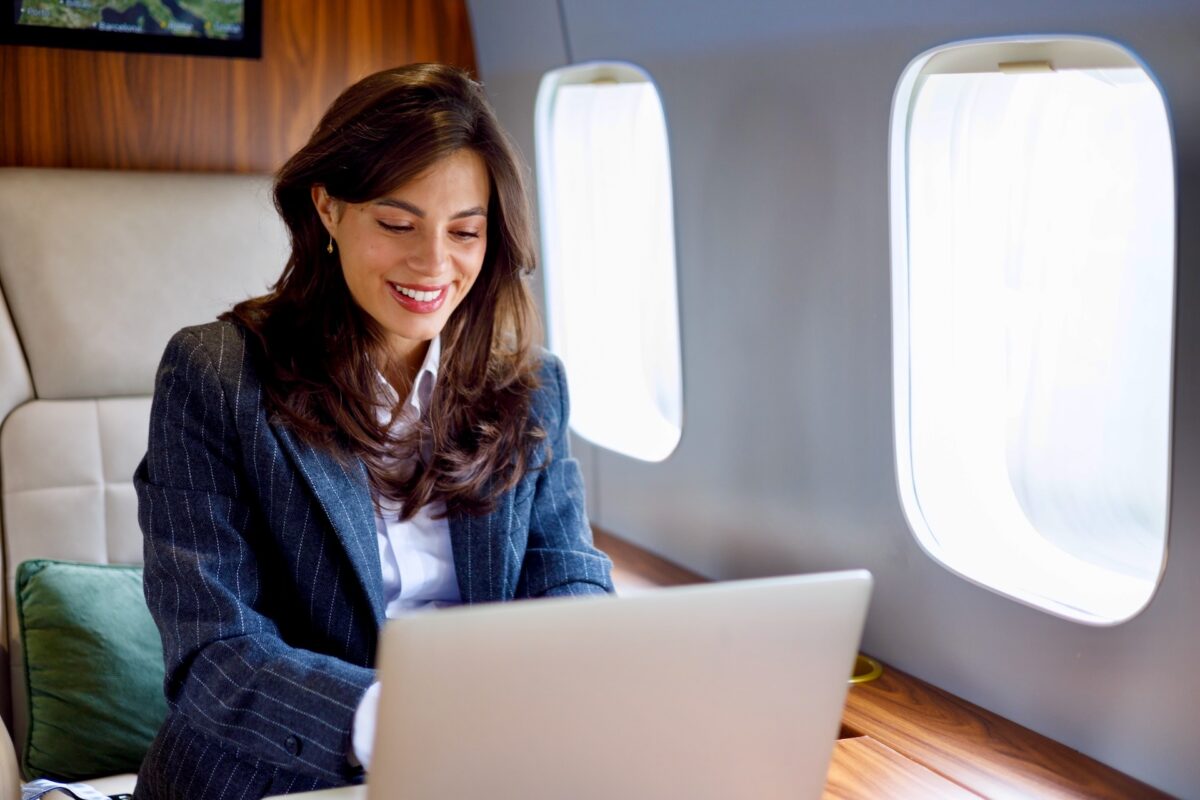Recently Saved 1 of 1 4 Businesswoman in a pinstripe blazer sits aboard a private jet smiling while using a laptop