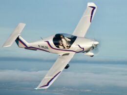 White aerobatic plane in a banked turn over the coast two people visible in the cockpit with flight suits on tail number F HLIX