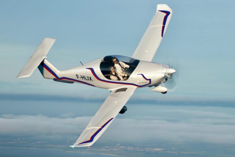 White aerobatic plane in a banked turn over the coast two people visible in the cockpit with flight suits on tail number F HLIX