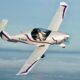 White aerobatic plane in a banked turn over the coast two people visible in the cockpit with flight suits on tail number F HLIX