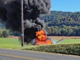 A small aircraft on fire in a rural field black smoke billowing and orange flames rising near a roadside hedge with wooded hills in the background