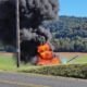 A small aircraft on fire in a rural field black smoke billowing and orange flames rising near a roadside hedge with wooded hills in the background