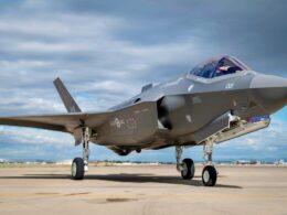 Side view of a gray stealth fighter jet on a tarmac cockpit canopy closed landing gear down in bright daylight under a cloudy sky