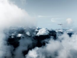 Airplane flying above a snow capped mountain range with clouds and clear blue sky ahead of the peaks