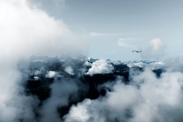 Airplane flying above a snow capped mountain range with clouds and clear blue sky ahead of the peaks