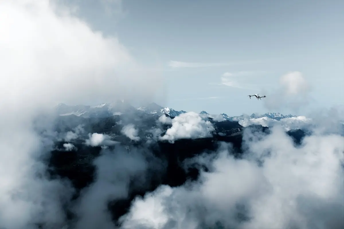 Airplane flying above a snow capped mountain range with clouds and clear blue sky ahead of the peaks
