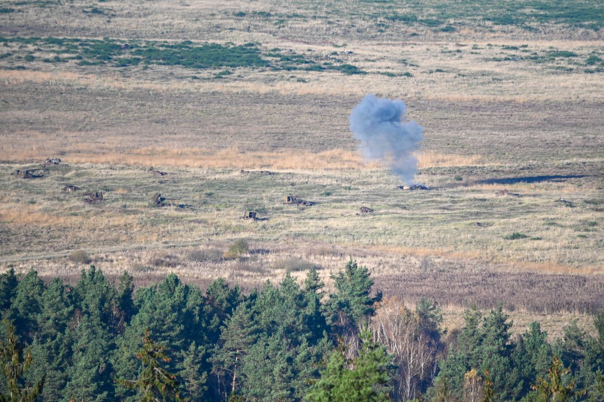 Switchblade 600 Loitering Munitions Training Open field with scattered military vehicles and a blue gray smoke plume rising from an explosion near center right with a forest edge in the foreground