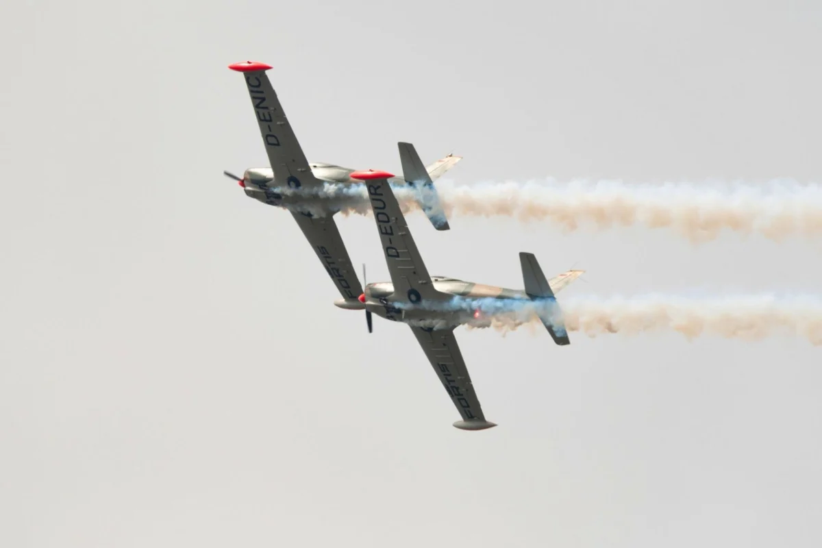 Philippines probes SF 260 collision that killed four pilots Two aerobatic airplanes flying in close formation emitting smoke trails against a pale sky one inverted showing red nose caps