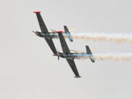 Two aerobatic airplanes flying in close formation emitting smoke trails against a pale sky one inverted showing red nose caps