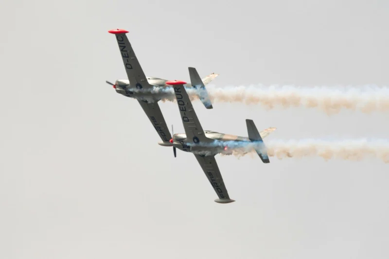 Two aerobatic airplanes flying in close formation emitting smoke trails against a pale sky one inverted showing red nose caps