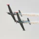 Two aerobatic airplanes flying in close formation emitting smoke trails against a pale sky one inverted showing red nose caps