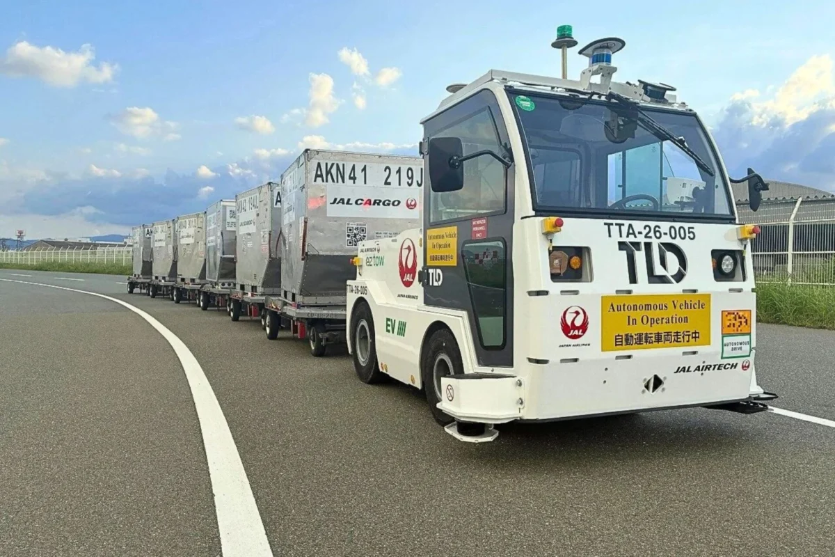 JAL brings driverless towing tractors to Fukuoka Airport Autonomous cargo vehicle pulling a line of silver shipping containers on a curved highway The truck has operation stickers and beacon lights on top