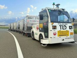 Autonomous cargo vehicle pulling a line of silver shipping containers on a curved highway The truck has operation stickers and beacon lights on top